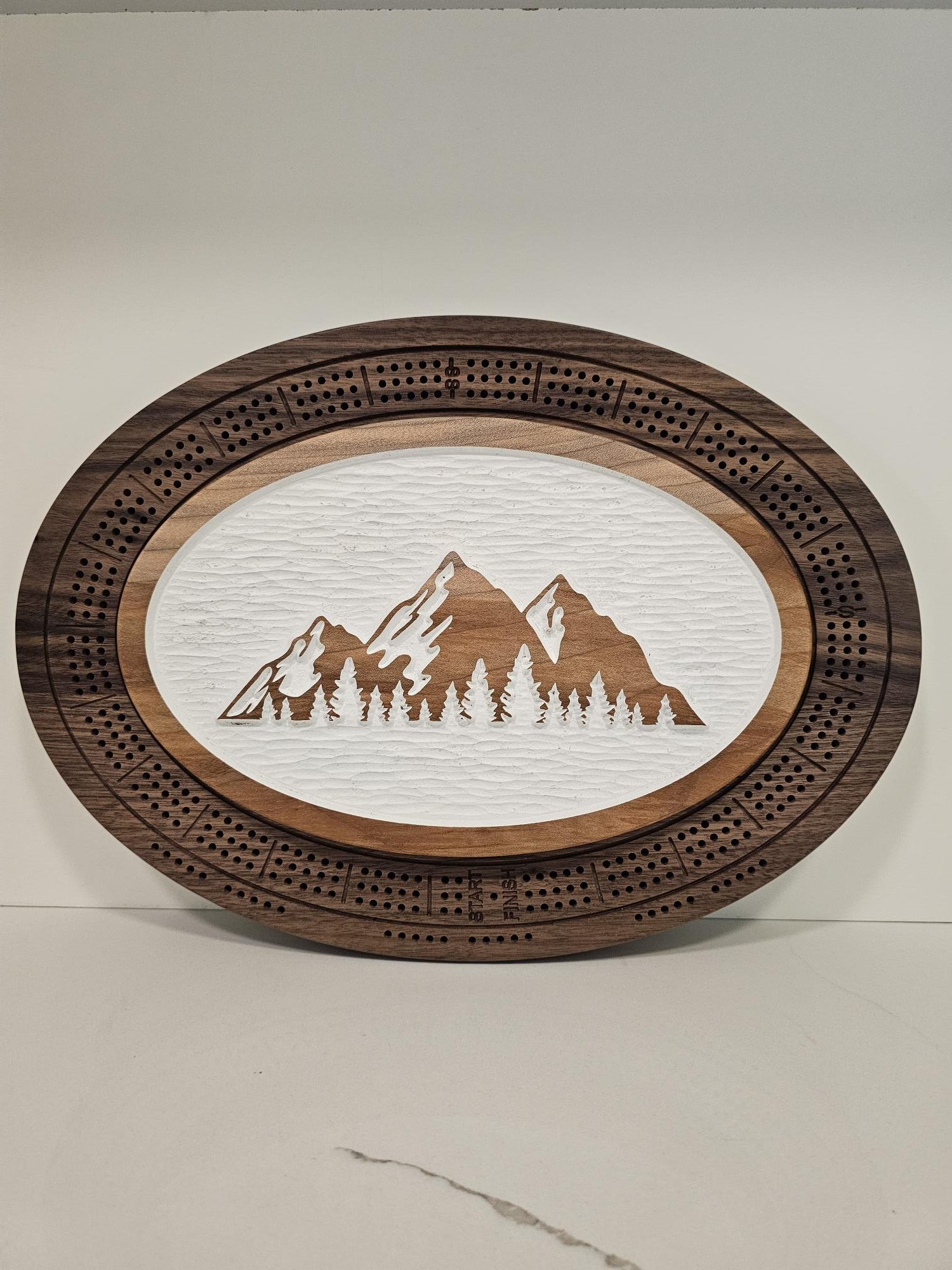 Oval wooden cribbage board featuring mountain and forest scene with natural wood grain background