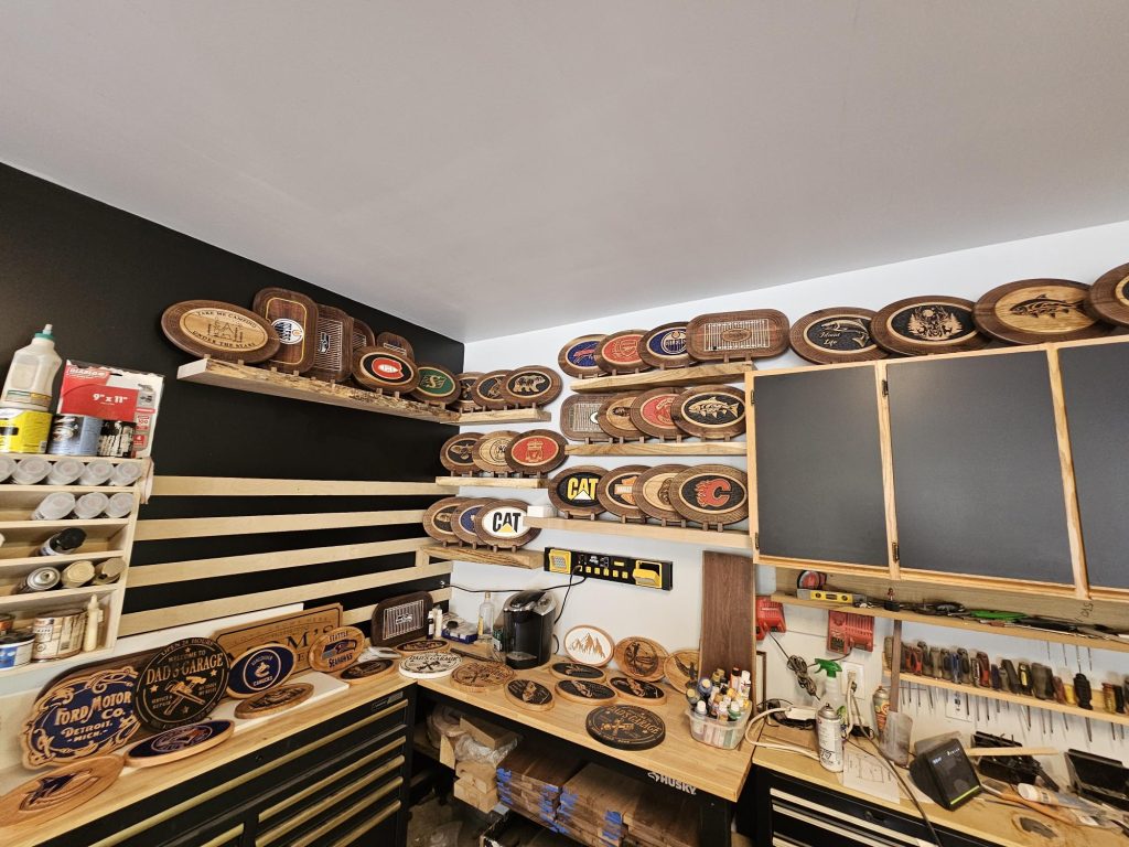 Workshop displaying custom wooden cribbage boards and signs on tiered shelving with tools and supplies